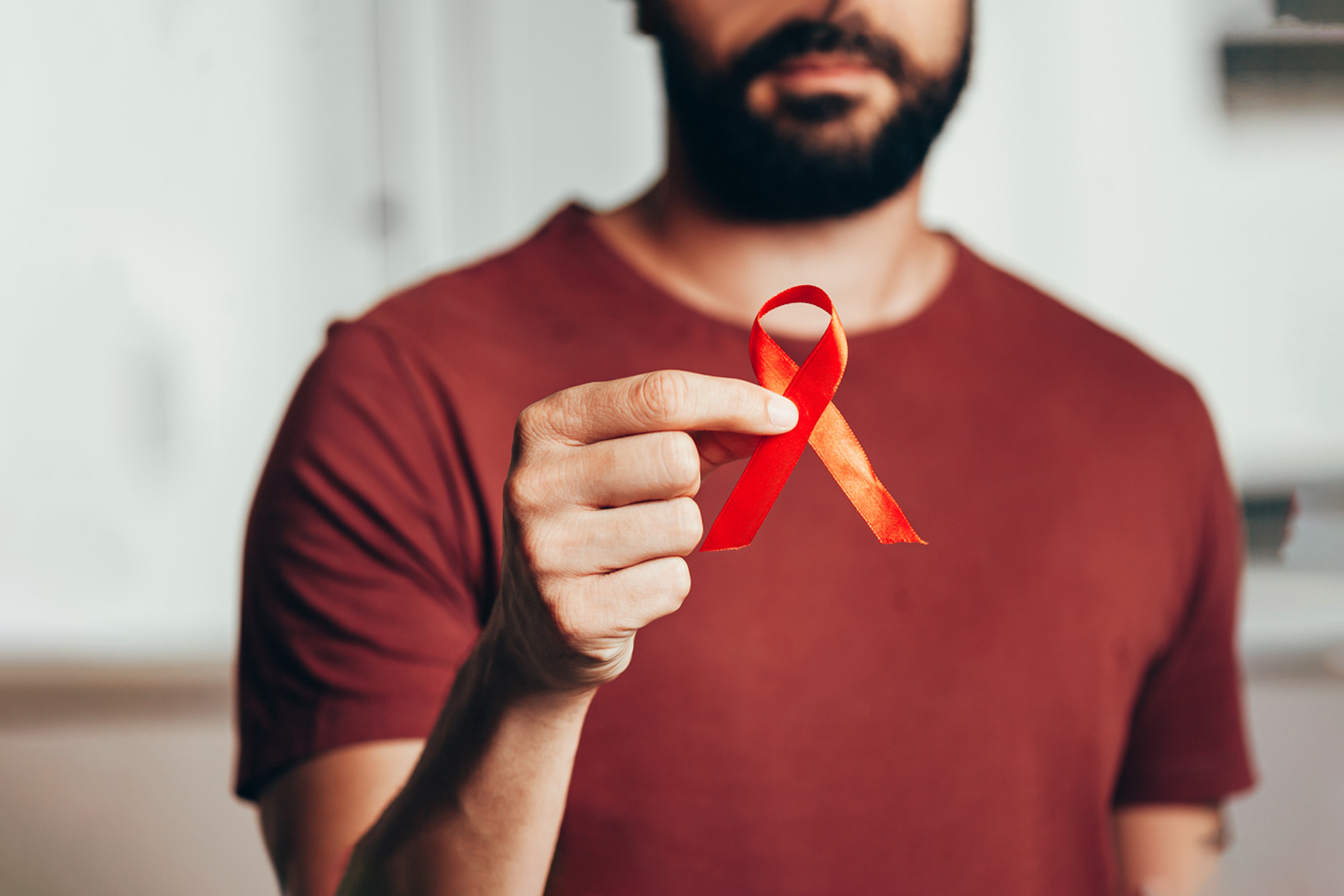 A bearded man in a red t-shirt holds a red AIDS ribbon
