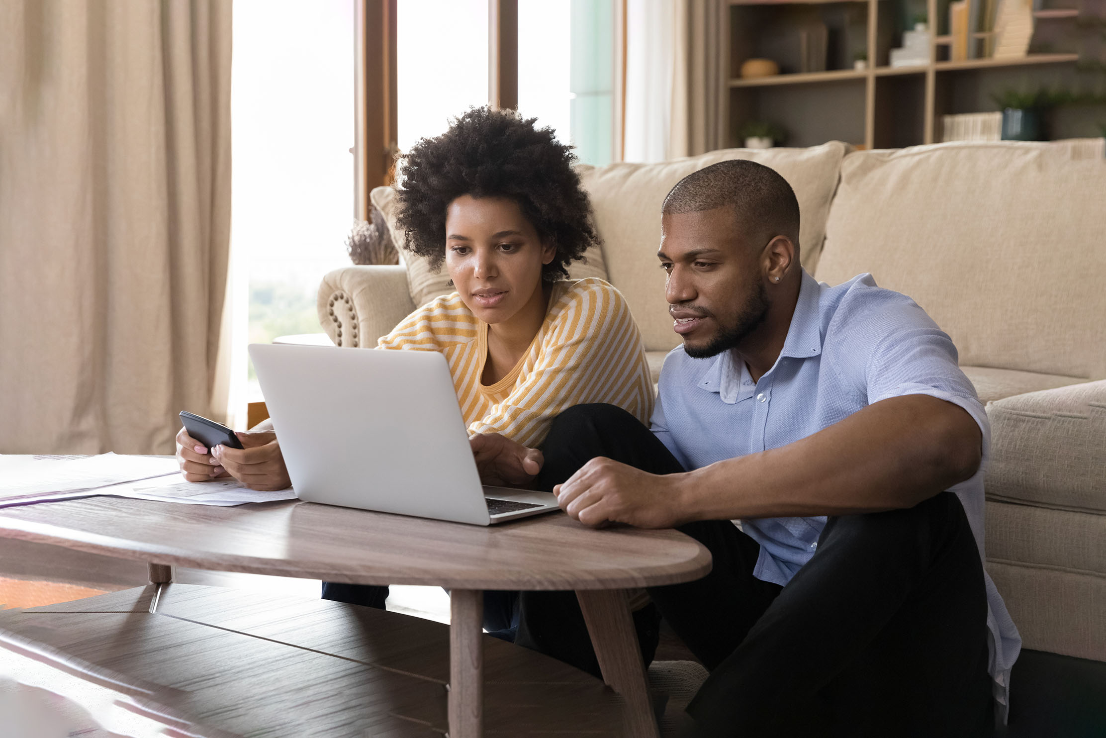 A black couple sit in front of the sofa, checking a laptop
