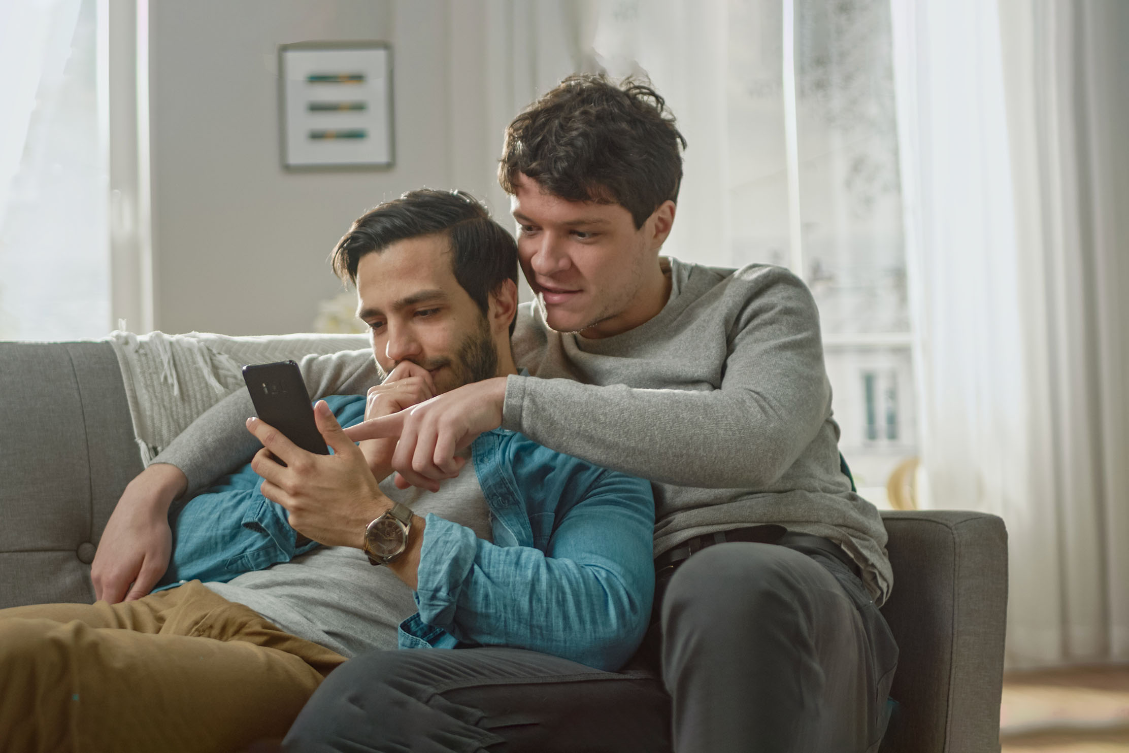 A same sex couple look at a mobile phone whilst sat on a sofa