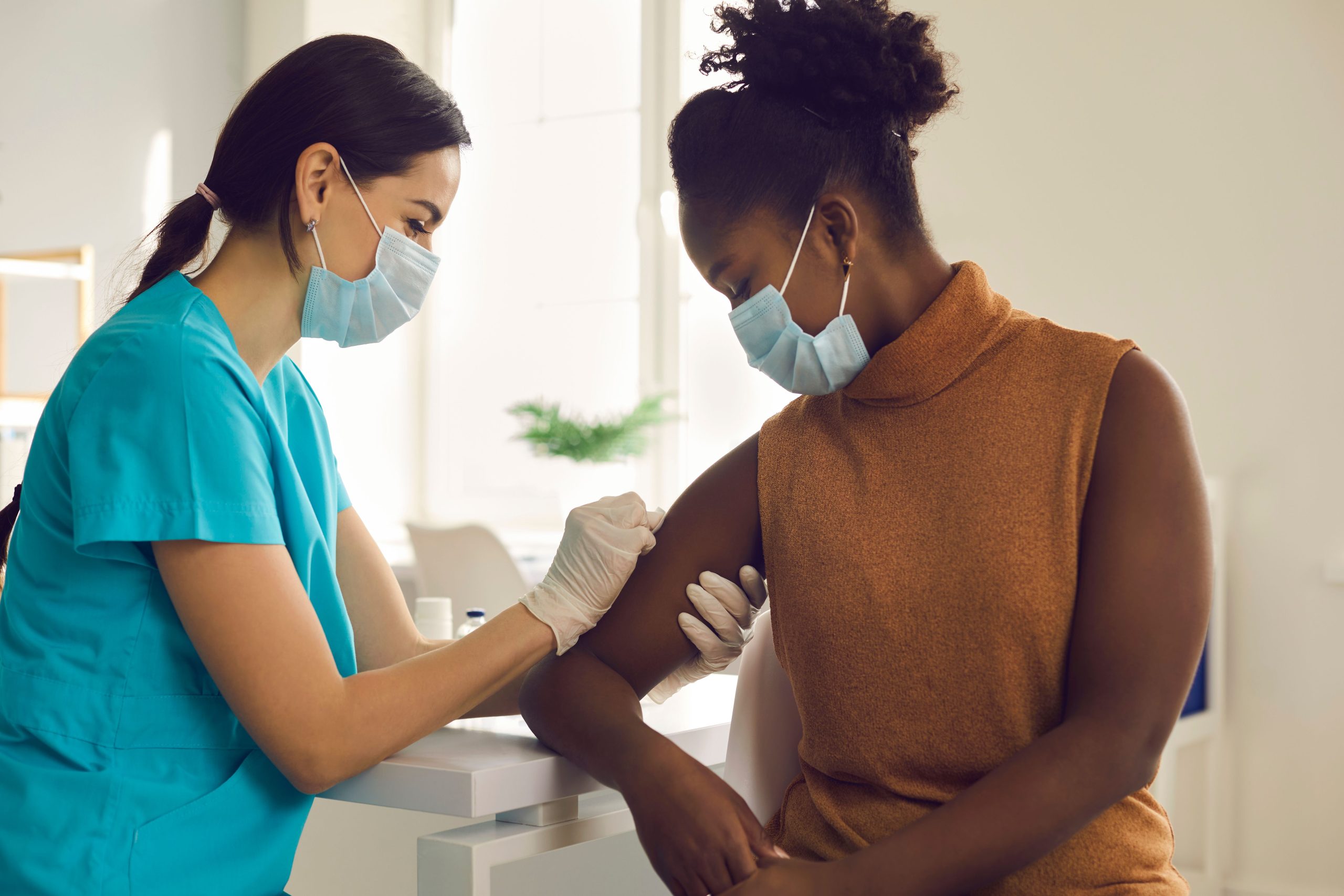 white nurse in green scrubs cleaning the arm of a black patient wearing a tan top preparing the injection point for a vaccine