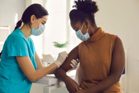 white nurse in green scrubs cleaning the arm of a black patient wearing a tan top preparing the injection point for a vaccine